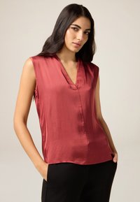 Ärmelloses Oberteil aus tiefrotem Satin mit einem V-Ausschnitt, lockerer Passform und glatter Textur. Kombiniert mit schwarzen Hosen, die ein modernes Design hervorheben.