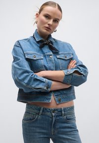 Μπλε τζιν μπουφάν με δύο τσέπες στο στήθος και κούμπωμα με κουμπιά, σε συνδυασμό με μπλε τζιν παντελόνι. Μαύρο μαντήλι γύρω από τον λαιμό προσθέτει αντίθεση.