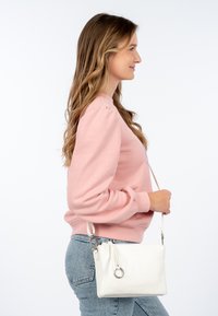 Bolso bandolera de cuero blanco con superficie texturizada, cierre de cremallera y detalle de anillo plateado. La modelo lleva un sudadera rosa y jeans.