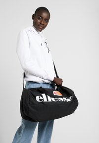 Svart duffelbag med stor hvit "ellesse"-logo, oransje detaljer, stoffhåndtak og justerbar skulderrem, vist holdt av en person.