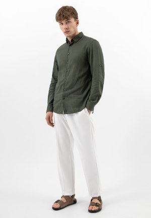 Jonge man staand, gekleed in een olijfgroen overhemd met knopen, witte broek en bruine sandalen met open tenen, tegen een effen witte achtergrond.