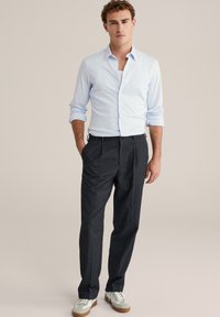 Lichtblauw overhemd met knopen, lange mouwen opgerold, gecombineerd met donkergrijze, op maat gemaakte broek en witte sneakers met grijze accenten.