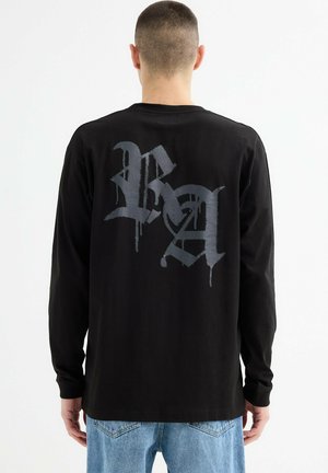 Hombre vistiendo una camisa de manga larga negra con letras góticas grises en la espalda, emparejada con jeans de mezclilla azul claros, de pie contra un fondo blanco.
