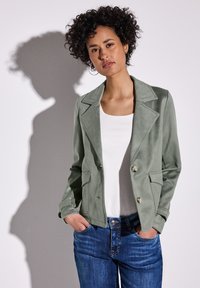 Hellgrüner, wildlederähnlicher, doppelt gefütterter Blazer mit Reverskragen, zwei Fronttaschen und silberfarbenen Knöpfen, kombiniert mit einem weißen Oberteil und blauen Jeans.