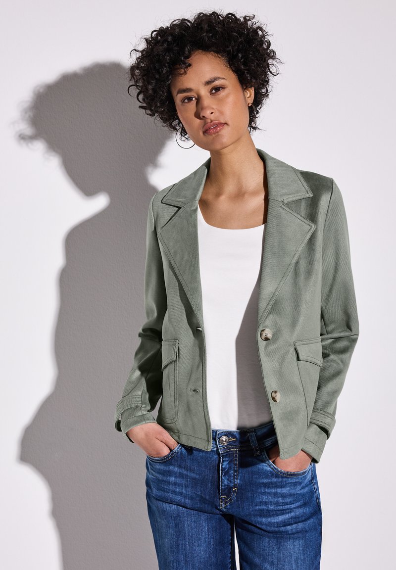 Hellgrüner, wildlederähnlicher, doppelt gefütterter Blazer mit Reverskragen, zwei Fronttaschen und silberfarbenen Knöpfen, kombiniert mit einem weißen Oberteil und blauen Jeans.
