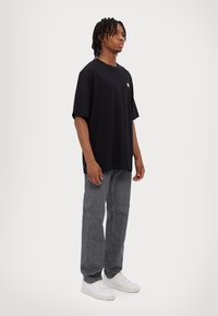 Black cotton oversized t-shirt featuring a small pink logo, paired with gray straight-leg jeans and white sneakers on a model.