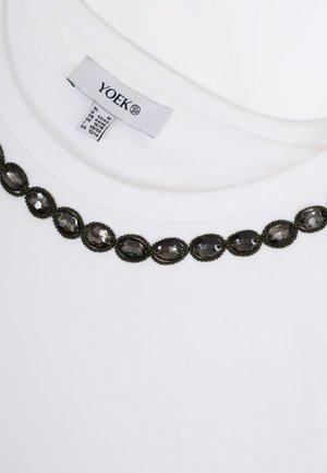 Encolure de vêtement blanc ornée d'embellissements noirs en forme d'ovale avec des bijoux, et une étiquette de vêtement visible indiquant "YOEK" avec les détails de la taille.