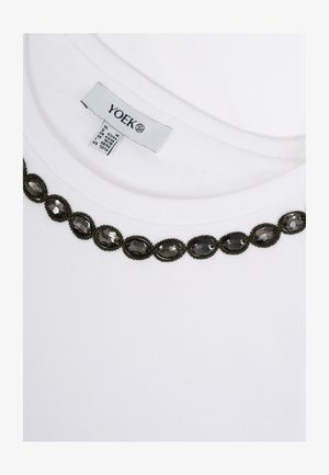 Weißer Kleidungs-Ausschnitt mit schwarzen, oval geformten, mit Juwelen besetzten Verzierungen und einem sichtbaren Kleidungsetikett mit der Aufschrift "YOEK" sowie Größenangaben.