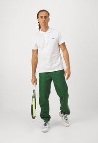 Polo bianco con logo verde Lacoste, abbinato a pantaloni verdi. I pantaloni presentano un design con strisce laterali. Tiene in mano una racchetta da tennis.