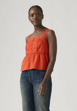 Kobieta o ciemnej karnacji, nosząca duże koła kolczyków, pomarańczowy top bez rękawów z guzikami oraz niebieskie dżinsy, stoi na szarym tle.