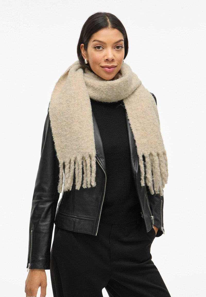 Écharpe beige douce avec des extrémités frangées, portée sur un pull noir et une veste en cuir. Texture douce dans une teinte chaude et neutre.