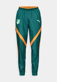 Pantalons de survêtement verts avec des rayures en chevron orange, une taille élastique et un patch logo sur la cuisse gauche. Fabriqués en tissu lisse.