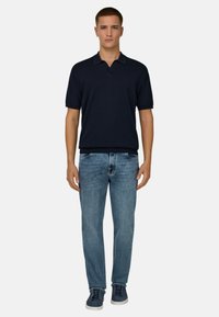 Jeune homme debout portant un polo noir à manches courtes, un jean bleu et des baskets bleu marine, sur un fond blanc uni.