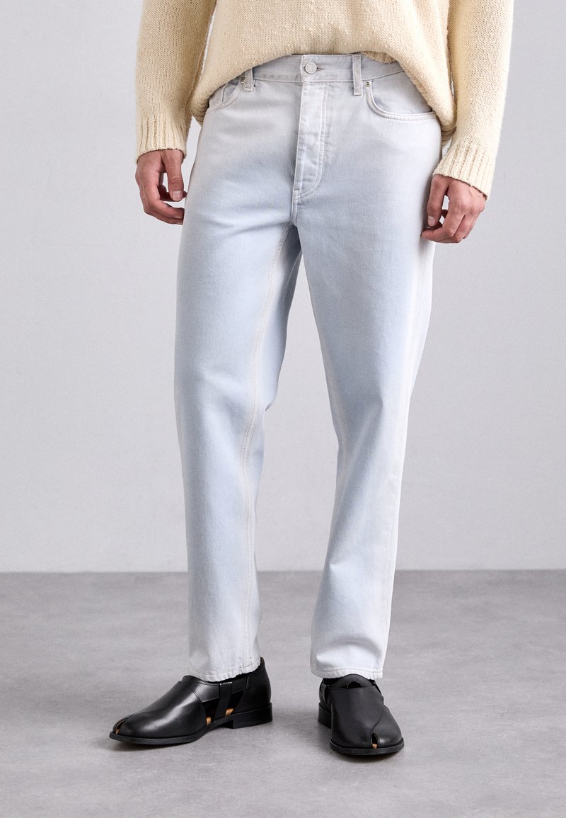 Light blue denim jeans with a straight cut, featuring visible stitching, side pockets, and a button closure, paired with black split-toe shoes.