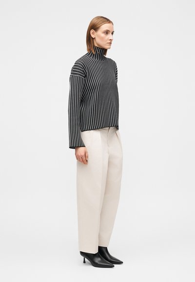 Πουλόβερ με ριγέ ζιβάγκο σε μαύρο και λευκό, συνδυασμένο με κρεμ παντελόνι φαρδιάς γραμμής. Το μοντέλο φοράει μαύρα μυτερά μποτάκια αστραγάλου.