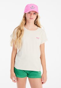 Camiseta de color beige claro con mangas cortas enrolladas, que presenta un pequeño logotipo "PROTEST" en rosa y verde; combinada con pantalones cortos verdes.