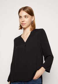 Blusa nera con scollo a V, maniche lunghe e una texture sottile. Caratterizzata da una vestibilità ampia e spacchi laterali, abbinata a jeans blu.