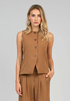 Femme blonde portant un gilet sans manches marron à boutons et un pantalon taille haute assorti, se tenant devant un fond gris uni.