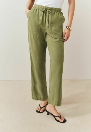 Persona che indossa pantaloni larghi verde oliva con grandi tasche frontali, top bianco, sandali neri con tacco e un grande braccialetto sul polso destro.