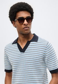 Άνδρας με κοντά σγουρά μαλλιά που φοράει σκούρα γυαλιά ηλίου και ένα πουκάμισο πόλο με κοντά μανίκια εμπριμέ, με μαύρο κολάρο.