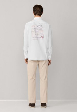 Hombre con camisa blanca de manga larga con un estampado colorido que dice "La Côte d'Azur 1950" en la espalda, pantalones beige y zapatos marrones, de pie mirando hacia la pared.