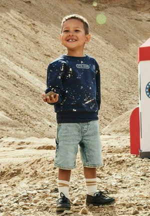 Lachende jongen in marineblauwe trui en denim short staat op rotsachtige grond en houdt een kleine steen vast bij een rood-wit speeltoestel.