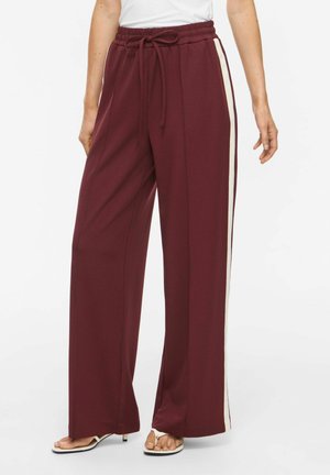 Pantalon bordeaux à jambes larges avec des bandes blanches sur les côtés, taille élastique avec cordon, porté avec des sandales blanches à talons hauts et un top blanc.