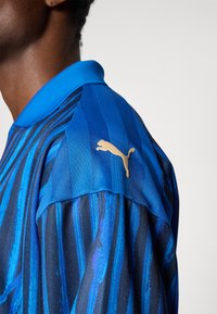 Plava prugasta polo majica s teksturiranim materijalom, koja sadrži zlatni Puma logo na ramenu. Dizajn s kragnom u kontrastnim bojama.