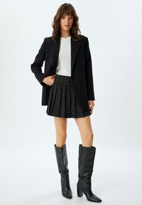 Blazer nero sopra un top bianco a coste, abbinato a una gonna plissettata nera con motivo a griglia bianca e stivali neri con tacco al ginocchio.