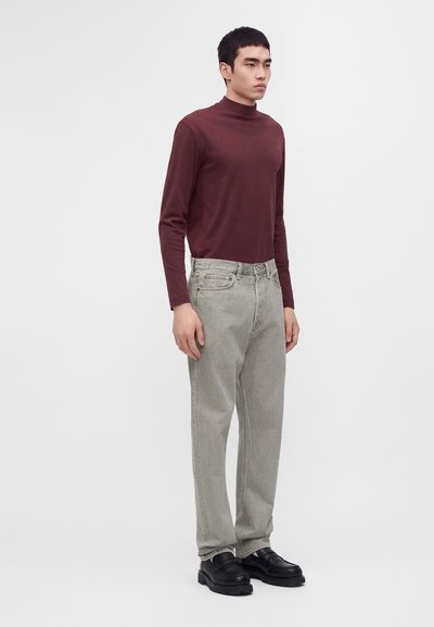 Jeune homme debout portant un col roulé bordeaux à manches longues, un jean gris clair et des chaussures noires épaisses sur un fond clair uni.
