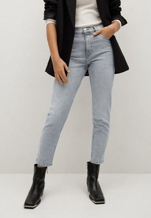 Femme portant un jean taille haute bleu clair, des bottines noires, un haut blanc côtelé et un blazer noir, les mains détendues et dans la poche.