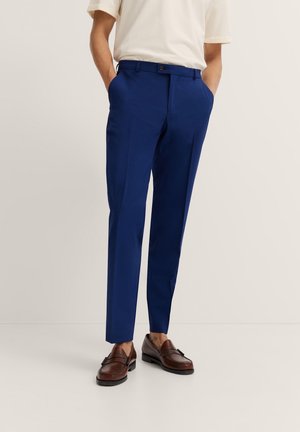Blaue Anzughosen mit glatter Textur und maßgeschneiderter Passform, ausgestattet mit Gürtelschlaufen, einem Knopfverschluss und Seitentaschen. Kombiniert mit braunen Loafers.
