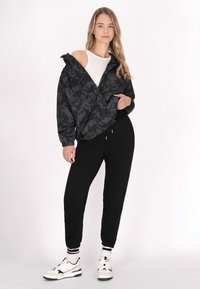 Sort mønstret jakke med elastiske manchetter, båret over en hvid top, parret med sorte joggerbukser og hvide sneakers med sorte accenter.