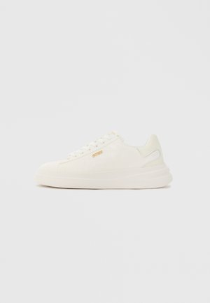 Zapatillas blancas con textura suave, patrón en relieve sutil, detalle de logo dorado y una suela gruesa y acolchada para mayor comodidad.