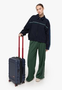 Marinblå rutig jacka med dragkedja, gröna vida byxor med vita ränder och en marinblå hård resväska med röd utdragbar handtag och hjul.