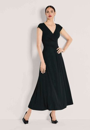 Frau in schwarzem, langem V-Ausschnitt-Kleid mit kurzen Ärmeln und schwarzen spitzen High Heels, die mit einer Hand auf der Hüfte vor hellem Hintergrund steht.