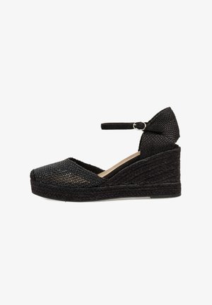 Espadrille negra con cuña, superior de malla tejida, tira en el tobillo y suela texturizada similar al yute. Presenta punta cerrada y parte trasera elevada.