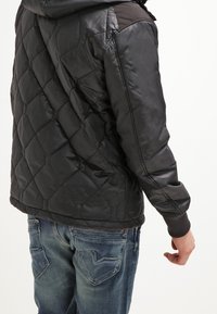 Svart vadderad jacka med huva, med diamantstickning, slät textur och ribbade ärmar; bärs med urblekta blå jeans.