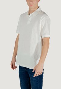 Polo shirt bianco a maniche corte con un sottile motivo testurizzato, una patta con due bottoni e dettagli a contrasto sul colletto. Abbinato a jeans blu.
