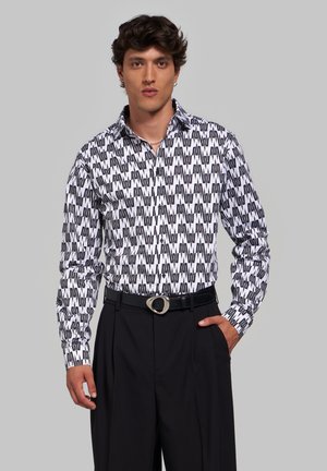 Homme portant une chemise blanche à manches longues avec un motif en zigzag noir et un pantalon noir taille haute, debout avec une main dans la poche.