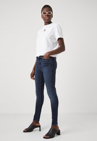 Camiseta blanca de manga corta con logo, vaqueros ajustados de color azul oscuro y mule de tacón bloque abierto en la parte delantera de color negro. Modelo de pie en perfil.