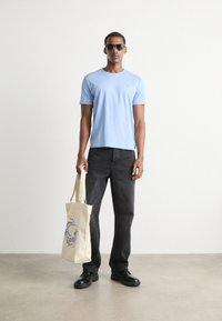 Light blue cotton t-shirt, black denim jeans, black shoes, and a cream tote bag with blue graphic design. Model wearing sunglasses.