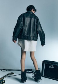 Giacca in pelle nera oversize, camicia bianca e stivali neri con frange alla caviglia. Sullo sfondo si vedono una chitarra e un amplificatore.