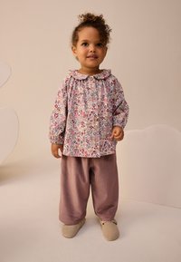 Világos rózsaszín virágmintás felső kerek gallérral és gombos előlappal. Laza, bordázott mályva nadrág, puha bézs cipővel párosítva.