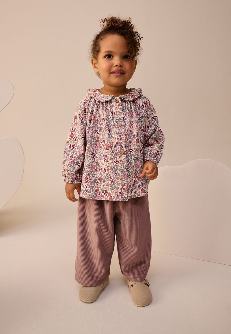 Haut à motif floral en rose pâle, avec un col rond et une patte de boutonnage à l'avant. Pantalon ample côtelé mauve, associé à des chaussures beige clair.
