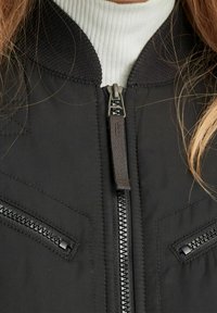 Czarna kurtka z przodem na zamek i dwoma zapinanymi kieszeniami, noszona na białym, prążkowanym swetrze z golfem.