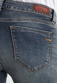 Tmavomodré džíny z denimu s zadní kapsou a oranžovým prošíváním a koženým štítkem s embossed nápisem "LTB." Hladká textura s mírným vyblednutím.