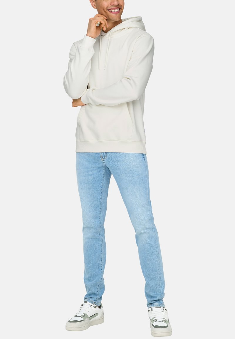 Haut à capuche beige avec une poche avant et un cordon de serrage, associé à un jean bleu clair et des baskets blanches avec des accents verts.