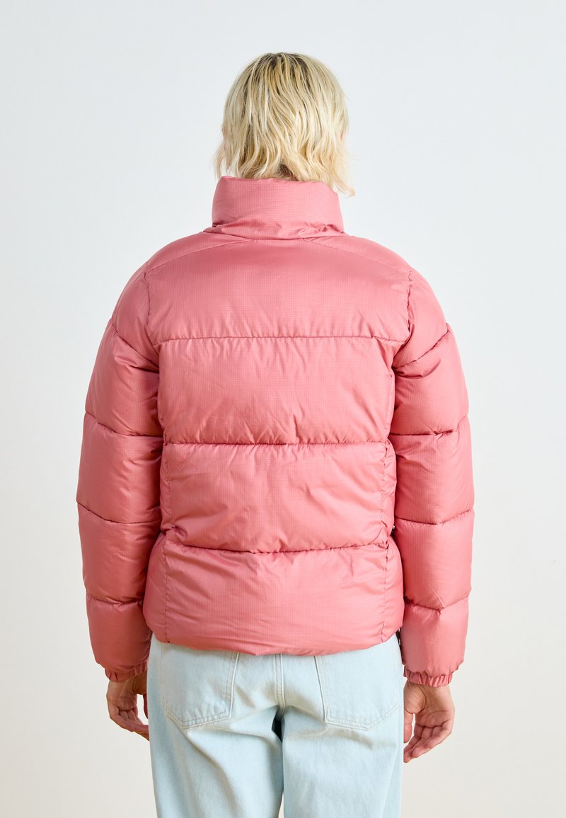 Veste en duvet rose avec un col haut, texture matelassée, poignets élastiques et une finition légèrement brillante. Portée sur un jean bleu clair.