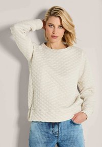 Blonde vrouw met blauwe ogen, gekleed in een gestikt cr�èmekleurig sweater en een blauwe spijkerbroek, poserend met één hand achter haar hoofd en de andere in een zak.